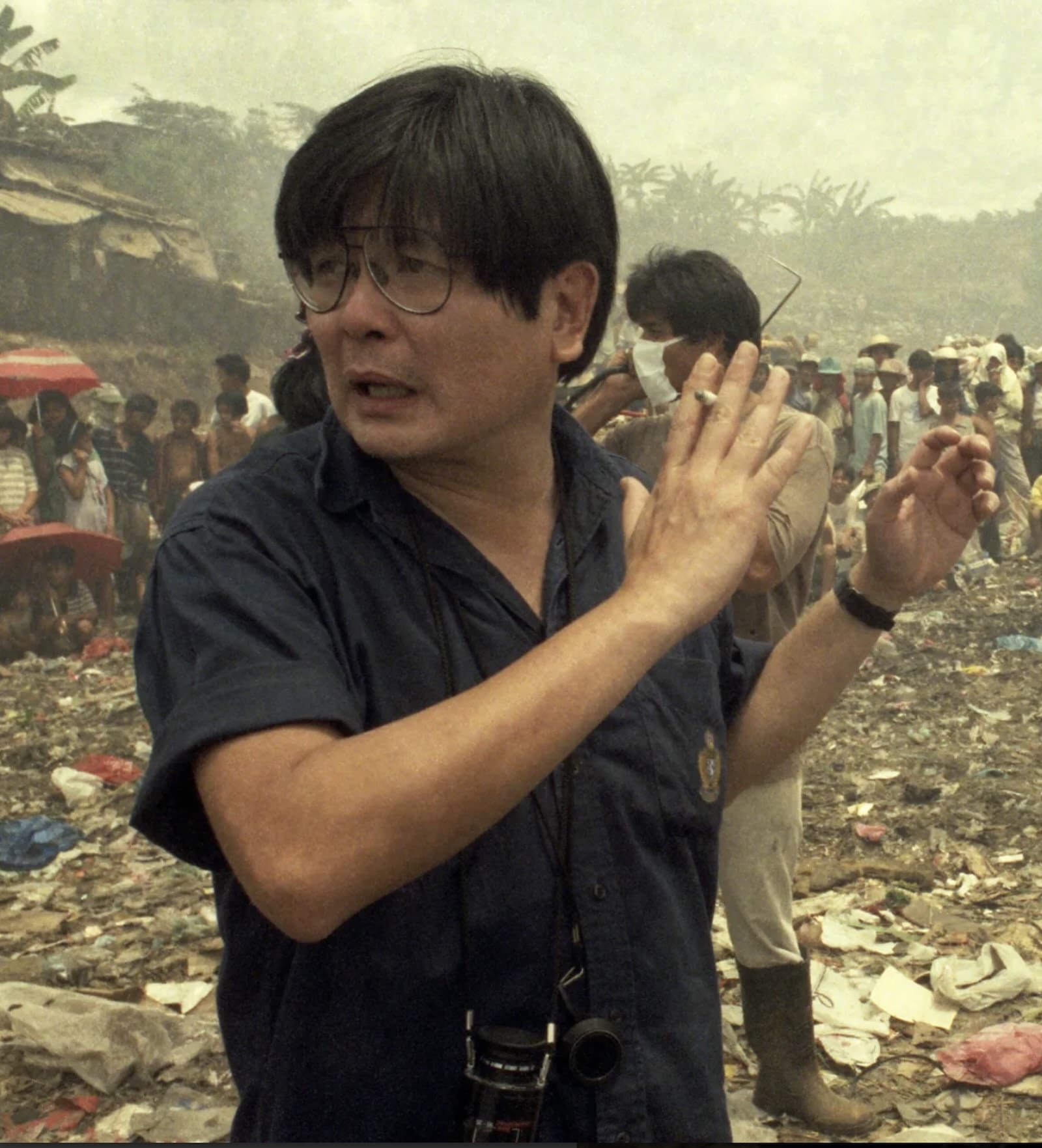 A man wearing a camera around his neck and holding a cigarette between his fingers stands in a crowded movie set